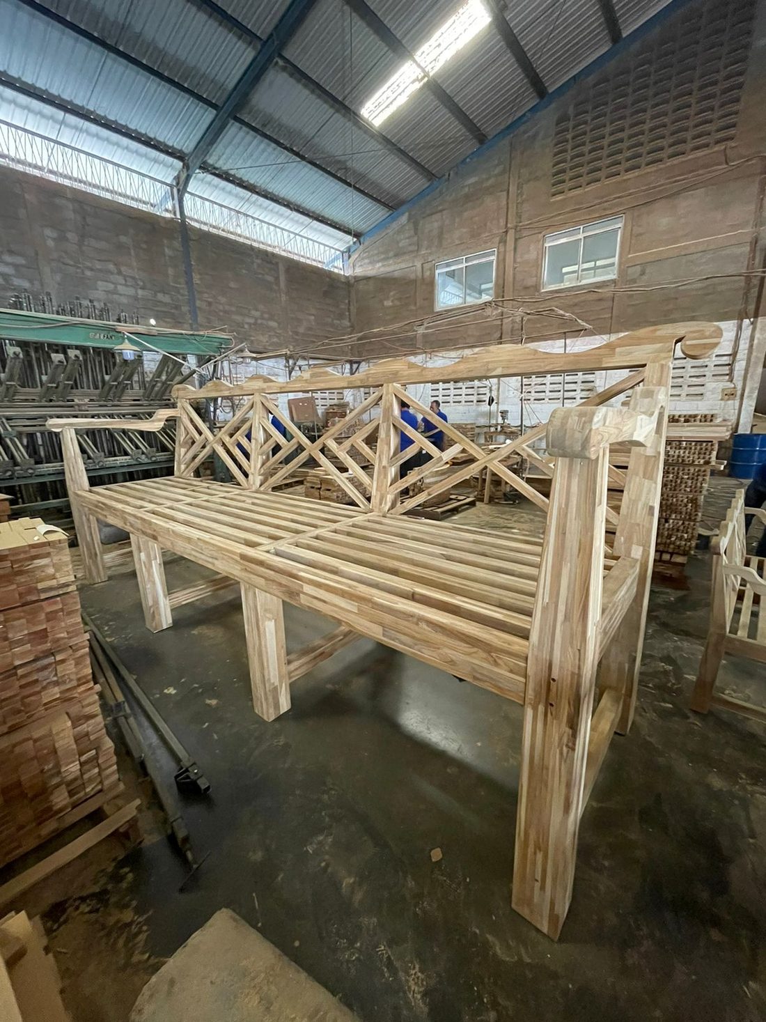 Eine dreisitzige Bank aus unbehandeltem, hellem Teak steht fertig montiert mitten in einer Werkstatt. Dahinter liegen Stapel von Teakrohlingen und halbfertigen Möbeln.