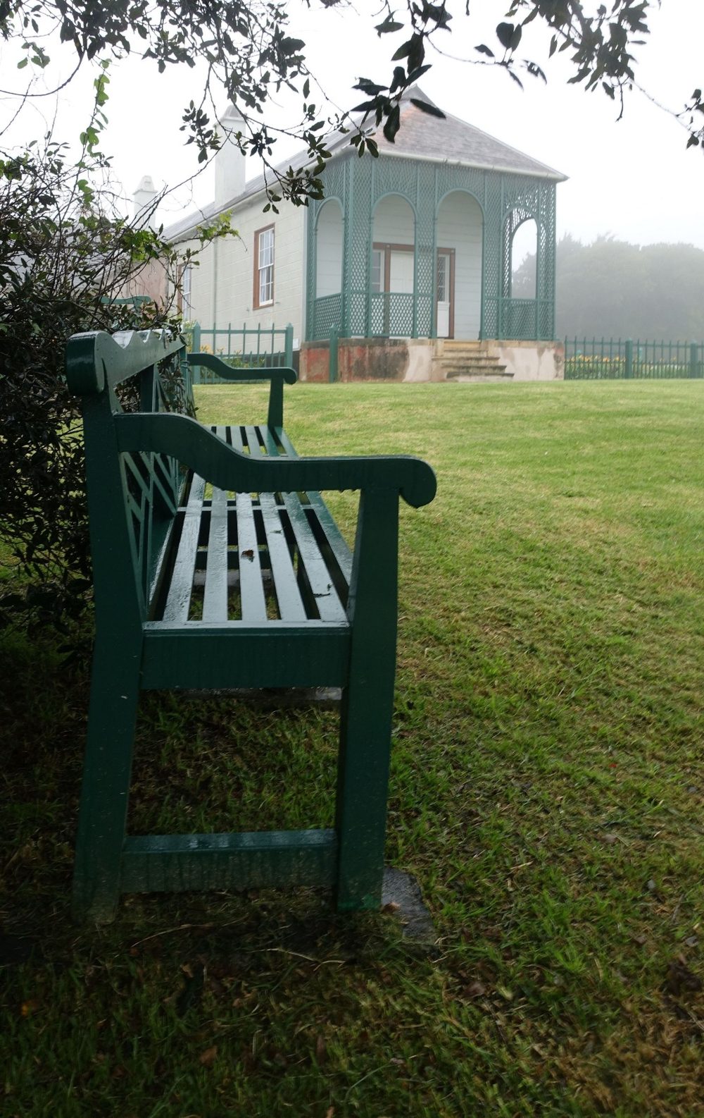 Die grüne Bank steht im Gras unter einem Baum bei Longwood House auf St. Helena. Dahinter liegt die grüne Veranda des Hauses im Nebel.