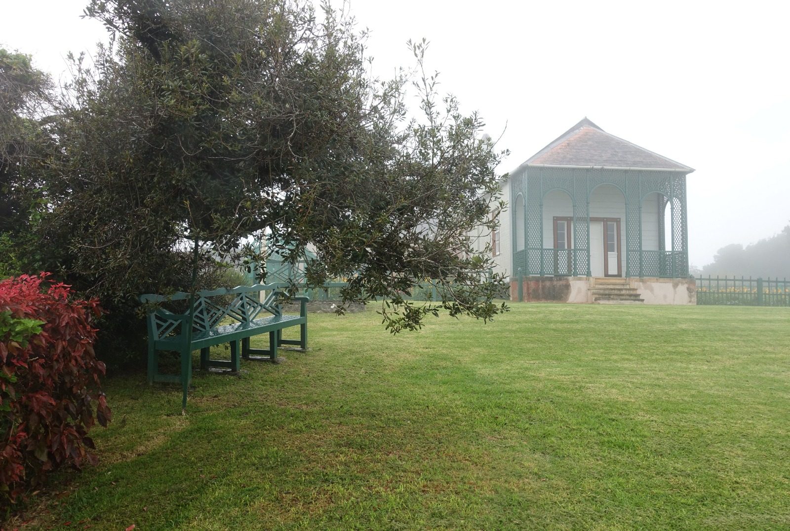 Longwood House über den Rasen hinweg im Nebel. Links steht die Bank im Schatten eines Baumes neben einem rotlaubigen Strauch.