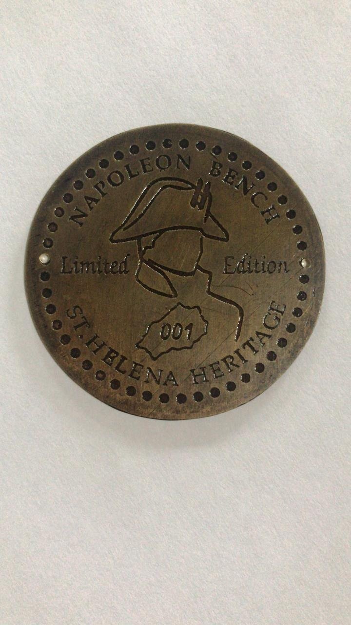 Rundes Messingschild mit Perlrand. Eingraviert ein Bicorne im Profil, der Text Napoleon Bench, Limited Edition, St. Helena Heritage und der Umriss der Insel mit der Nummer 001.